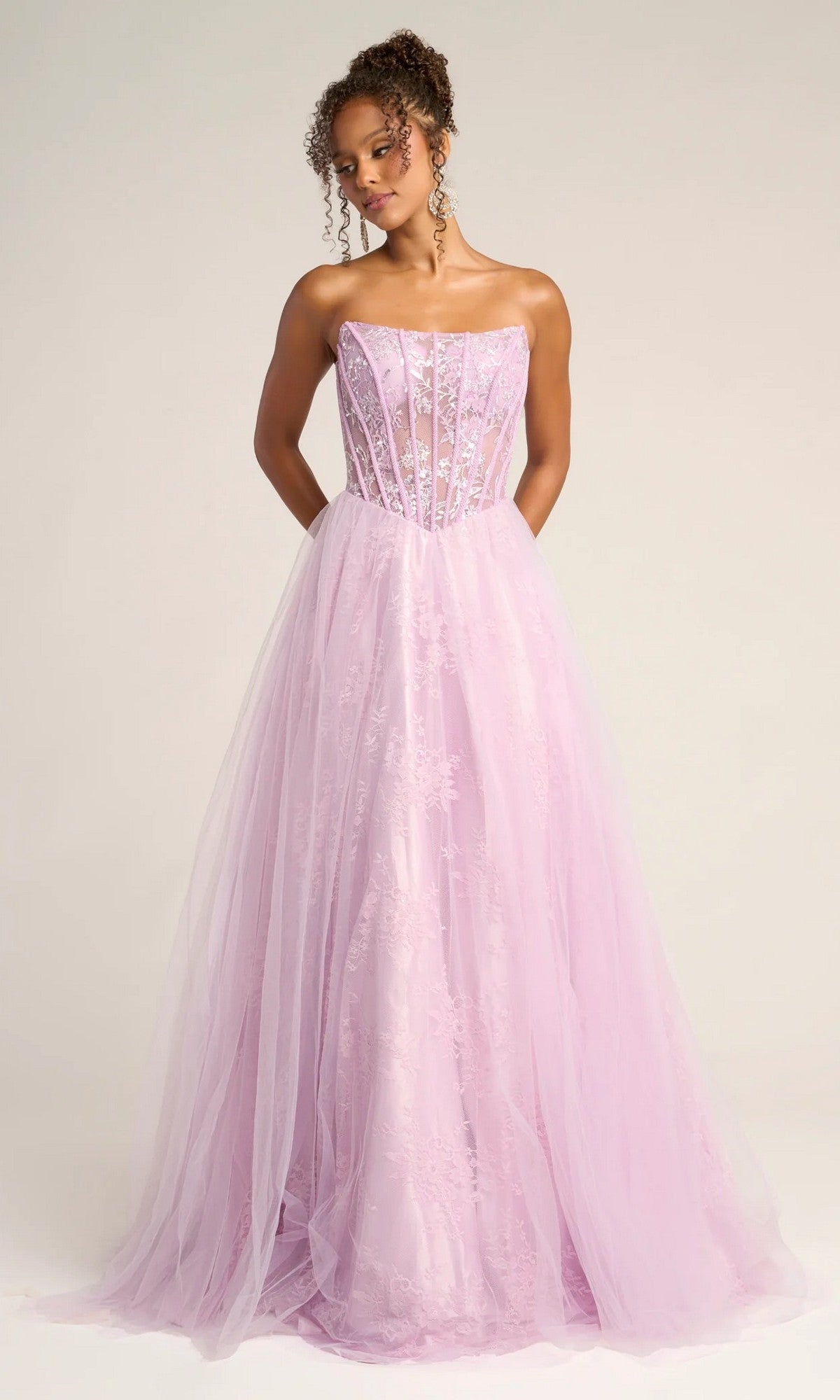 A woman stands against a plain background wearing the Portia and Scarlett XO023 long formal dress, featuring a strapless lace ball gown design with a sheer corset bodice and a flowing tulle and lace skirt.